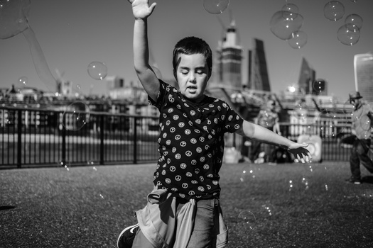 Young child blowing bubbles with city skyline in background, captured in black and white.