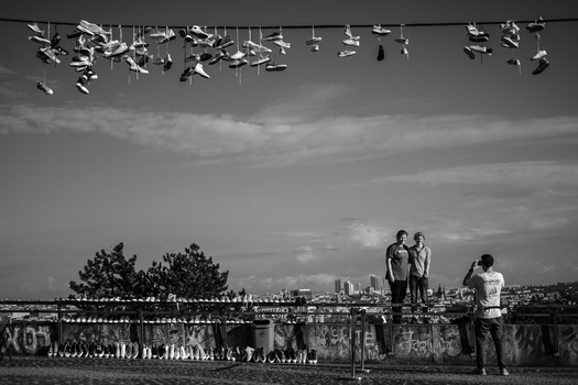 Two people standing on a rooftop with city skyline in background; shoes hanging from power line above.