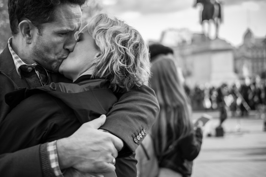 Couple kissing in an urban plaza while other people walk in the blurred background.