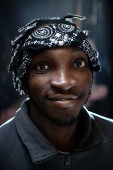 Portrait of a person wearing an ornate patterned head wrap, shot against a dark background with studio lighting.