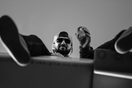 Bearded man wearing sunglasses and headphones, photographed from below against a gray sky.