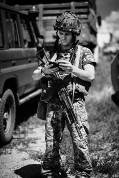 Soldier in camouflage uniform holding rifle while standing outdoors near a vehicle.