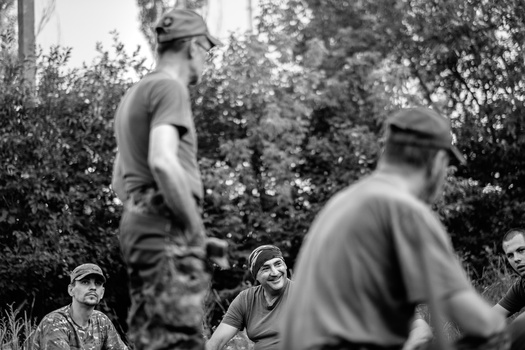 Black and white photograph of people gathered outdoors among trees, with selective focus on two seated figures in foreground.