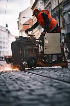 Street photographer in orange jacket adjusting camera on vintage suitcase amid cobblestone urban setting.