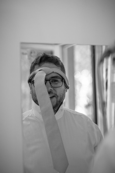 Man wearing glasses and white shirt adjusting eyeglasses while looking at camera in black and white portrait.