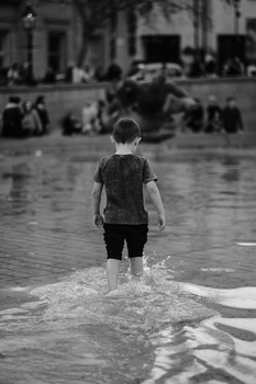 Child wading into shallow water in an urban plaza, photographed from behind in black and white.