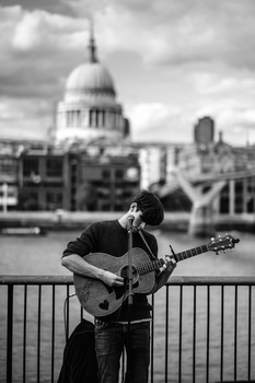 Musician playing electric guitar on a bridge with a domed cathedral visible across the water behind him.