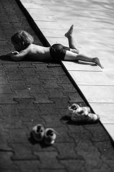 Child lying on wooden dock beside water with shoes in foreground, shot in black and white.