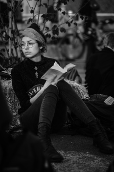 Woman wearing glasses and a beanie reads a book while seated outdoors, surrounded by blurred foliage.