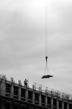 A wrecking ball hangs from a cable above a concrete building under demolition against an overcast sky.