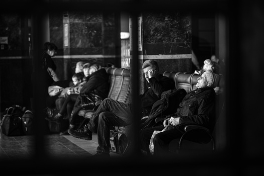 Black and white photograph of people waiting in what appears to be a transit station or public space, framed through a structural column.