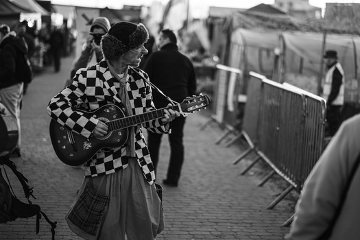Street musician playing acoustic guitar while wearing a checkered jacket and hat.