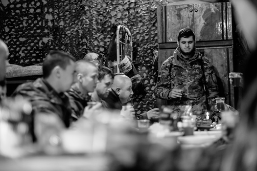 Soldier speaking to seated military personnel in a camouflaged tent setting.