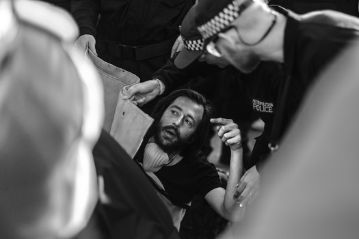 Bearded man surrounded by police officers during what appears to be an arrest or detention.