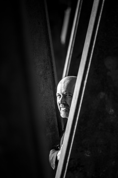 Bald man peering through vertical lines or bars in dramatic black and white portrait.