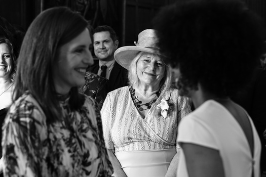 Black and white photograph of women conversing at an indoor gathering, with men visible in the background.