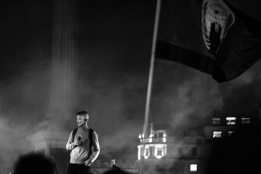 Performer on stage under dramatic spotlighting with flag banner overhead in black and white cinematography.