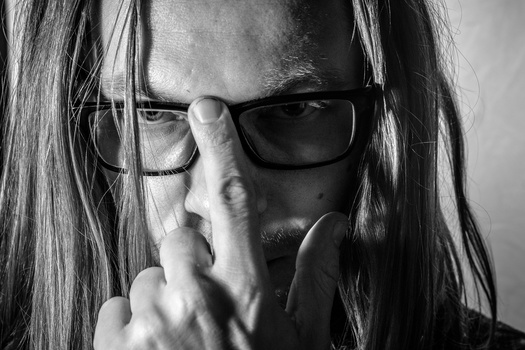 Person with long dark hair and glasses, finger to lips in a hushing gesture, shot in black and white.