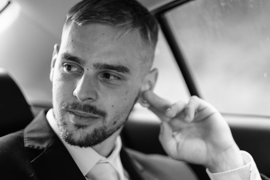 Black and white portrait of a man in a suit inside a car, looking toward the camera with hand raised.