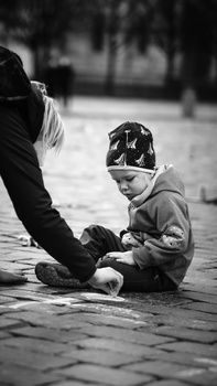 Young child sitting on pavement reaching toward an adult's extended hand in black and white.