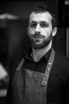 Black and white portrait of a bearded man wearing an apron, lit with dramatic side lighting against a dark background.