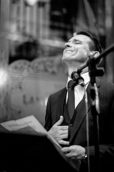 Man in suit and tie speaking at microphone, looking upward with a smile.