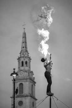 Performer on elevated platform shooting pyrotechnic flames skyward beside a church steeple.