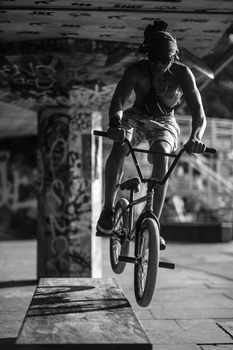 Rider performing a wheelie on a BMX bike under a concrete overpass in black and white.