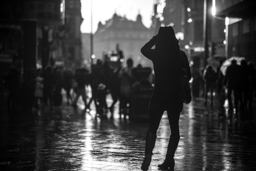 Silhouetted figure standing alone on a wet city street amid blurred crowds and architecture.