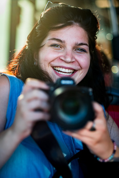Smiling person holding a DSLR camera with a telephoto lens, wearing blue and red clothing indoors.