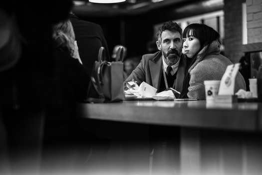 Black and white photograph of a man and woman sitting at a counter, with a blurred figure in the foreground.
