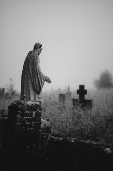 Draped stone angel statue standing on a grave in a misty cemetery with crosses visible in the background.