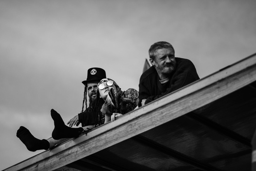 Two men sitting on a wooden roof beam against an overcast sky in black and white.