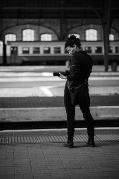 Person in dark jacket standing on brick platform in front of classical train station building, shot in black and white.
