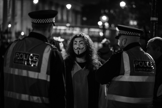 Person wearing Guy Fawkes mask surrounded by police officers at night on an urban street.