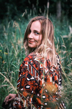 Young girl with long blonde hair wearing an ornate rust-colored patterned garment in a field.