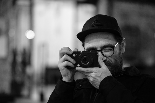 Bearded photographer wearing glasses and a black cap peers through a rangefinder camera.