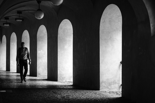 Solitary figure walking through a series of tall concrete archways in a stark black and white architectural space.