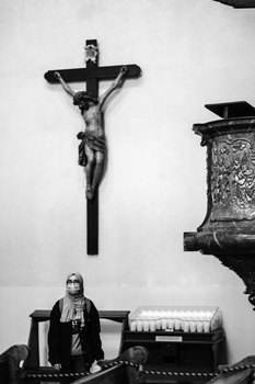 Black and white photograph of a crucifix mounted on a white wall above a seated priest in religious vestments.