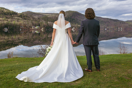 Bride and groom holding hands standing on grass overlooking a calm lake with forested mountains.