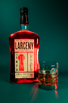 Bottle of Larceny bourbon whiskey next to a glass filled with amber liquid and ice against a teal background.
