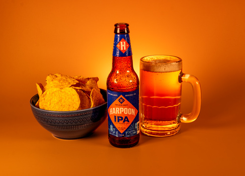 Corona IPA beer bottle next to a full glass mug and bowl of tortilla chips against warm orange background.
