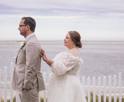 Bride and groom standing by white picket fence with ocean view backdrop.