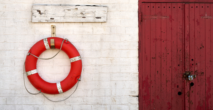 Red life preserver hung on white brick wall next to weathered wooden sign and red painted doors.
