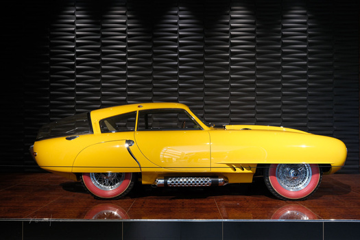 Yellow vintage car with red-wall tires displayed against a geometric textured wall backdrop.