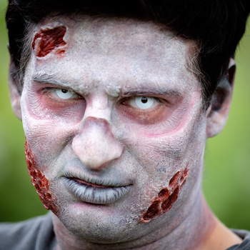 Close-up portrait of zombie character makeup with pale skin, bloodshot eyes, and prosthetic wounds on forehead and chin against green background.