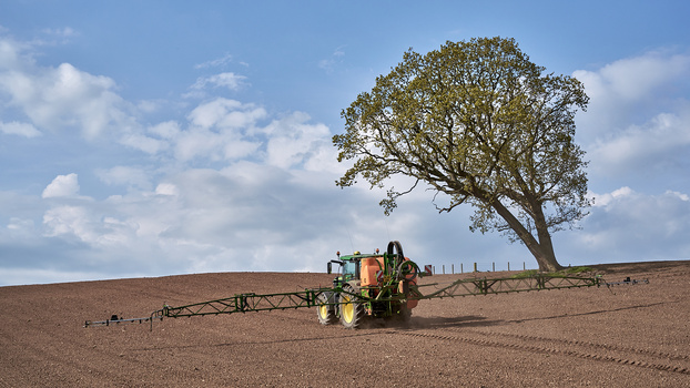 Green and yellow tractor with spray boom spraying an open field under a solitary tree and blue sky.
