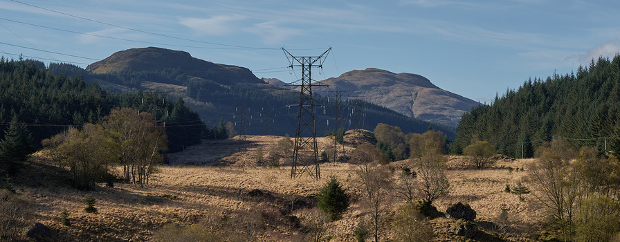 High-voltage transmission tower in valley with forested hills and mountains in background.
