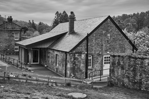 Stone cottage with slate roof and modern extension, photographed in black and white with forested hills in background.