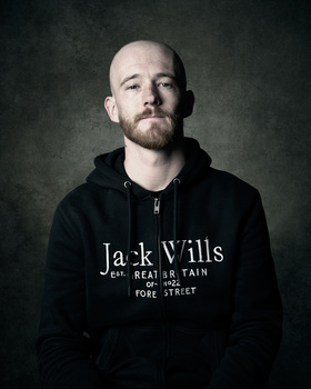 Portrait of a bald man with facial hair wearing a black hoodie against a textured dark background.